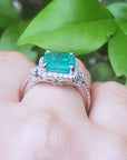 Emerald ring with diamond accents on a hand against a green leafy background.