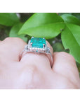 Emerald ring with diamond accents on a hand against a green leafy background.