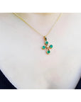 18K yellow gold cross pendant with four round Colombian emeralds and a small diamond, worn on a gold chain.