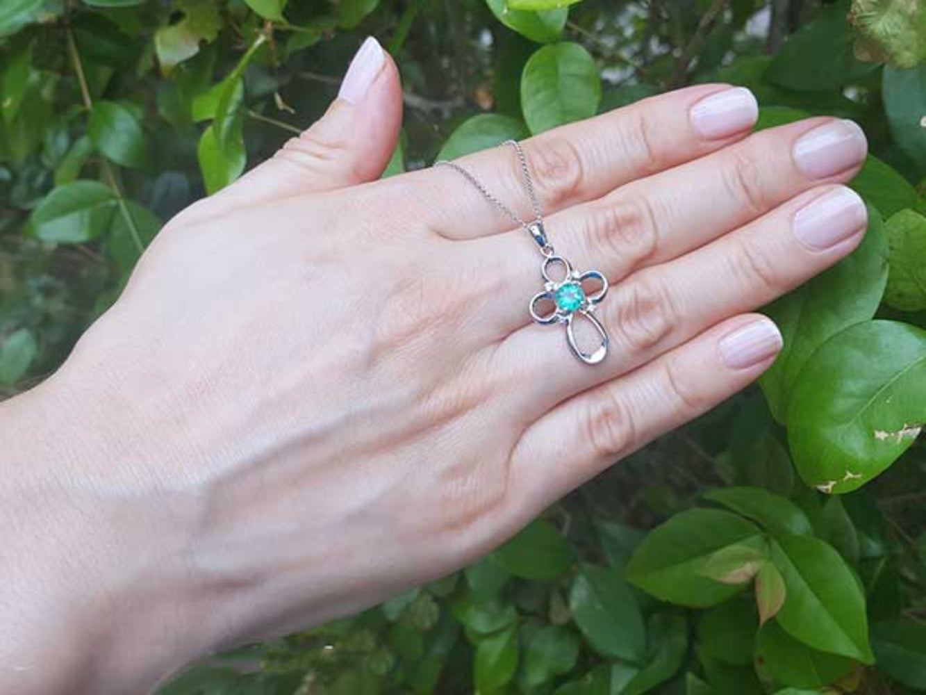 Hand holding a white gold necklace with a floral emerald pendant against a green leafy background.
