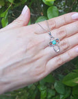 Hand holding a white gold necklace with a floral emerald pendant against a green leafy background.