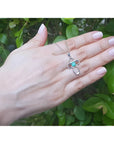 14K white gold cross pendant with a vibrant blue-green opal center, held by a hand against green leaves.