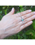 14K white gold cross pendant with a vibrant blue-green opal center, held by a hand against green leaves.