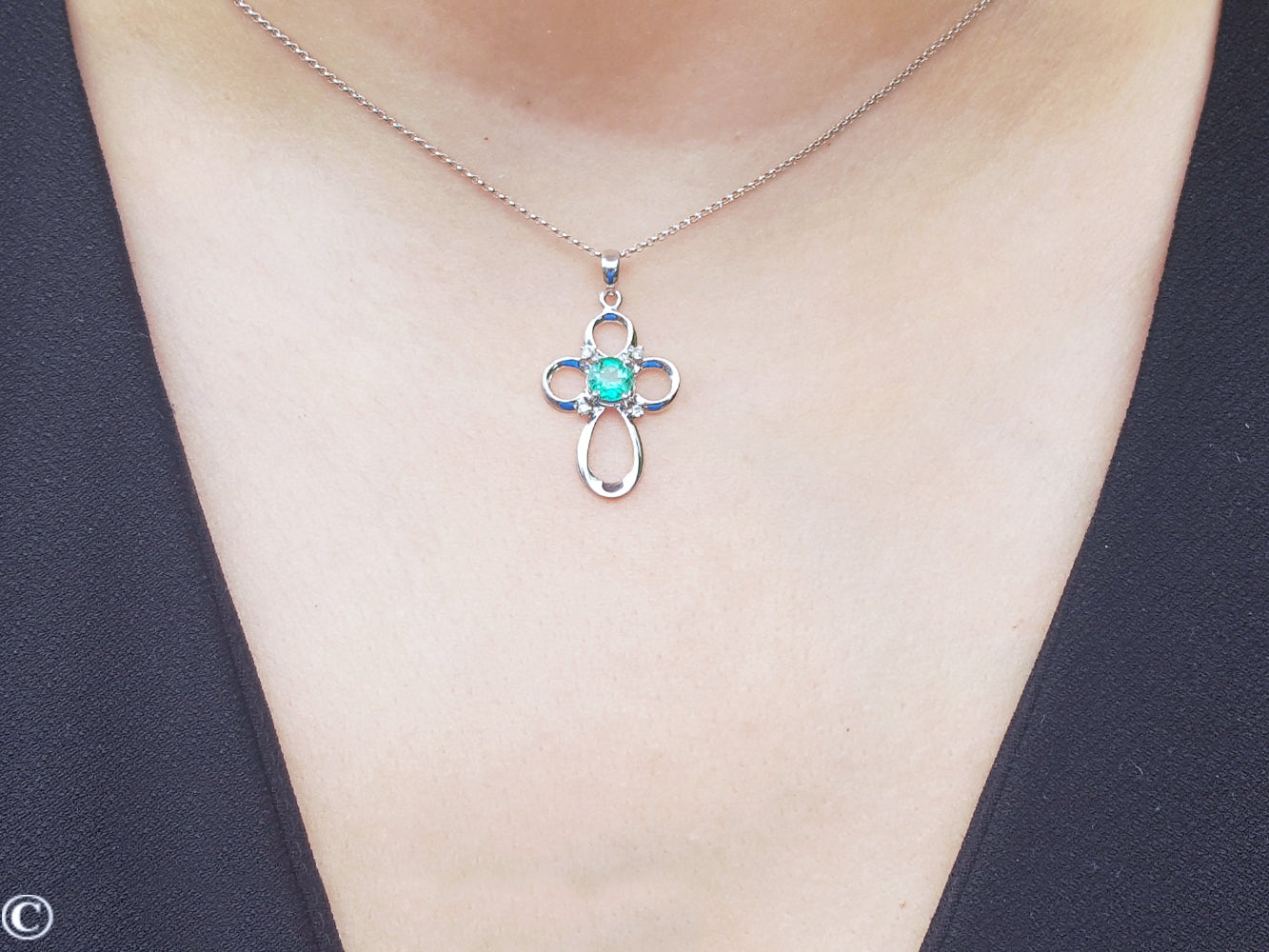 A white gold necklace with a round cut emerald pendant on a person wearing a black top.