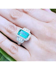 Woman's hand wearing an 18K white gold ring with a vibrant green emerald, halo, and baguette diamonds.