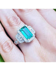 18K white gold ring with a vibrant green emerald, halo diamonds, and baguette side stones on a hand.