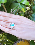 Hand wearing a Colombian emerald and diamond ring against a green leafy background.