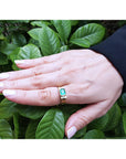 Hand wearing a gold ring for men with a natural emerald against a green leafy background.