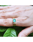 A yellow gold men's emerald ring on a hand against a leafy background.