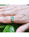 A yellow gold men's emerald ring on a hand against a leafy background.