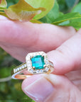 Gold ring with a square emerald and diamonds held by a hand against a green leafy background.