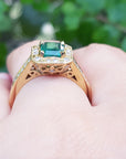 Gold ring with a natural emerald on a hand against a blurred green background.