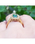 Gold ring with a natural emerald on a hand against a blurred green background.