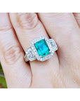 A natural emerald ring with diamond accents on a finger against a natural background.