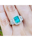 A natural emerald ring with a halo diamond accent on a finger is made of 18k white gold.
