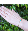 Hand wearing a gold bracelet with a emerald stone against a green leafy background.