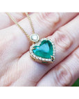 Heart-shaped emerald pendant with diamond halo on an 18k gold chain, held against a hand.
