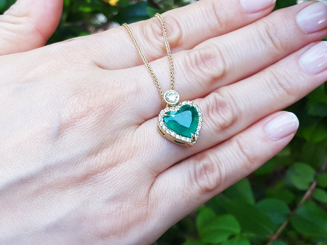 Hand holding a necklace with a emerald heart-shaped pendant against a blurred green background.
