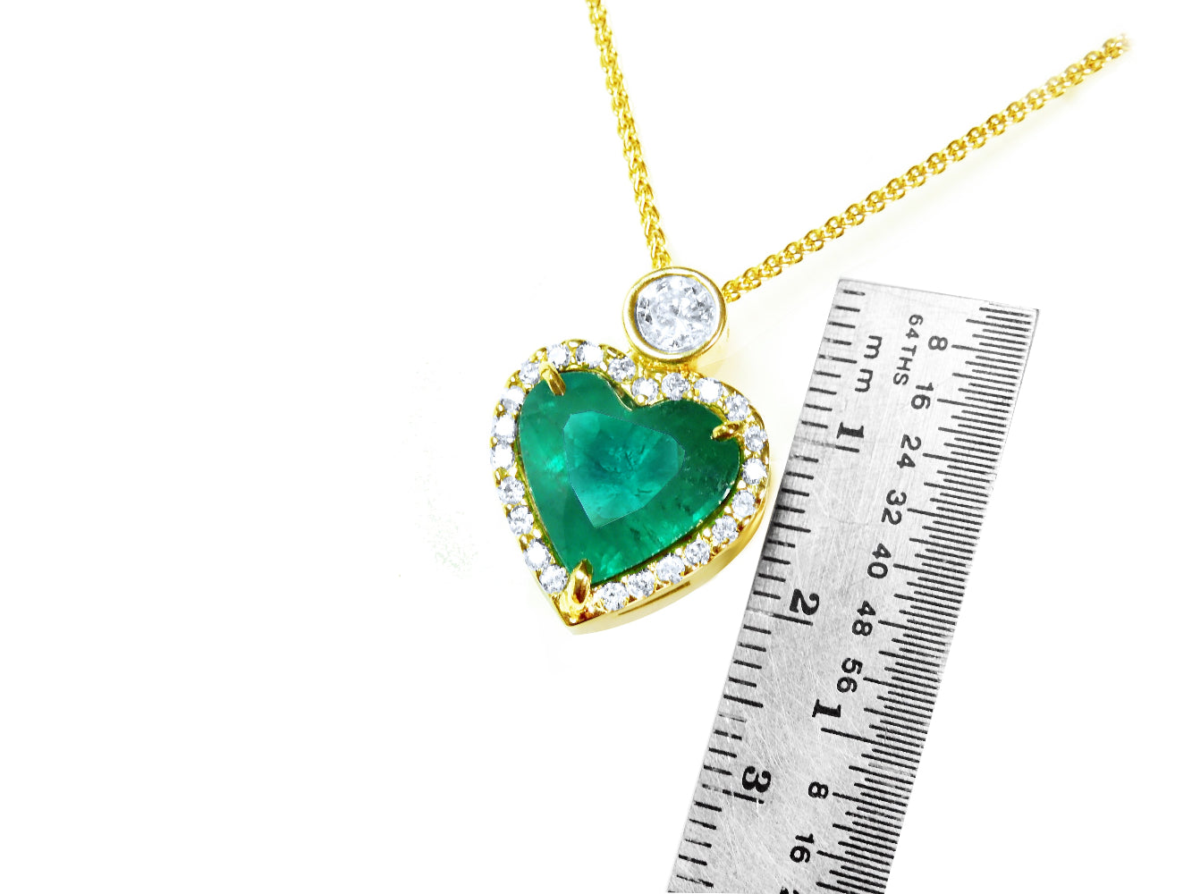 Heart-shaped emerald pendant with gold border and white diamond on top, next to a ruler for scale on a white background.