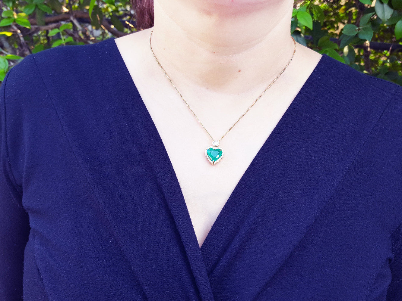 A woman wearing a blue blazer with a gold necklace featuring a natural emerald heart pendant.