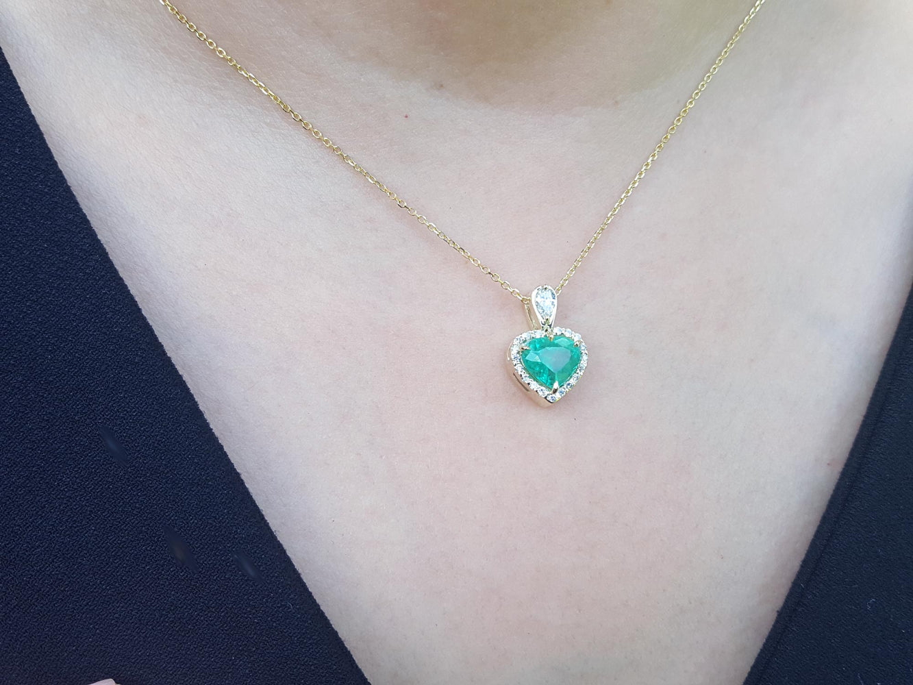 Gold necklace with a heart-shaped emerald pendant on a person wearing a dark top.