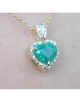 Heart-shaped emerald pendant with diamond accents on a gold chain against a light pink background