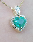 Heart-shaped emerald pendant with diamond accents on a gold chain against a light pink background
