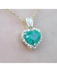 Heart-shaped emerald pendant with diamond accents on a gold chain against a light pink background