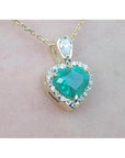 Heart-shaped emerald pendant with diamond accents on a gold chain against a light pink background