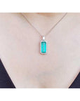 Elongated natural emerald pendant with halo diamonds on an 18K white gold chain, worn on a person.