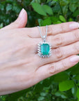 A 14k white gold emerald pendant on a person's hand against a natural green leafy background.