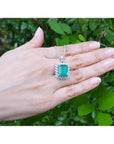 A 14k white gold emerald pendant on a person's hand against a natural green leafy background.
