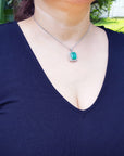 A woman wearing a navy blue dress with a deep neckline and a necklace with a natural emerald pendant, outdoors.