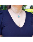 A woman wearing a navy blue dress with a deep neckline and a necklace with a natural emerald pendant, outdoors.