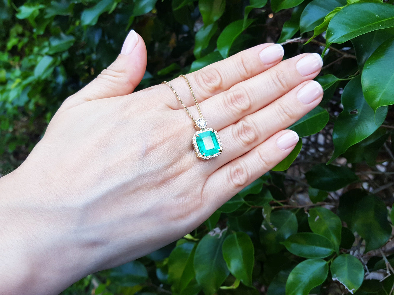 Hand displaying a square natural emerald pendant against a leafy background.