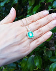 Hand displaying a square natural emerald pendant against a leafy background.