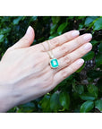 18K yellow gold pendant necklace with a rectangular emerald and diamond halo, held by a hand.