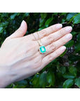18K yellow gold pendant necklace with a rectangular emerald and diamond halo, held by a hand.