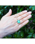 18K yellow gold pendant necklace with a rectangular emerald and diamond halo, held by a hand.