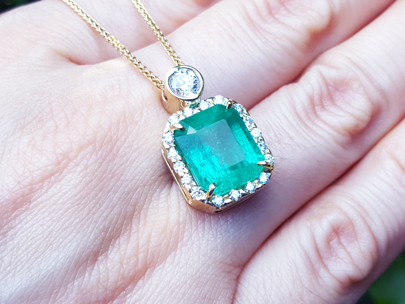 Emerald rectangular shaped pendant on a gold chain held on a hand.