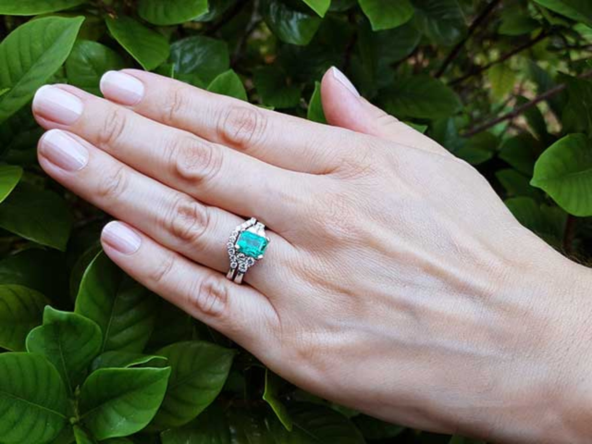 Hand wearing a natural emerald ring and matching band with green leaves in the background.