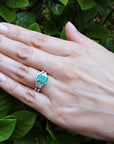 Hand wearing a natural emerald ring and matching band with green leaves in the background.