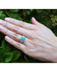 Hand wearing a natural emerald ring and matching band with green leaves in the background.