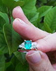 Hand holding a natural colombian emerald ring against a green leafy background.