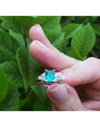Hand holding a natural colombian emerald ring against a green leafy background.