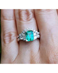 A real Colombian emerald and matching band ring on a finger against a natural background.