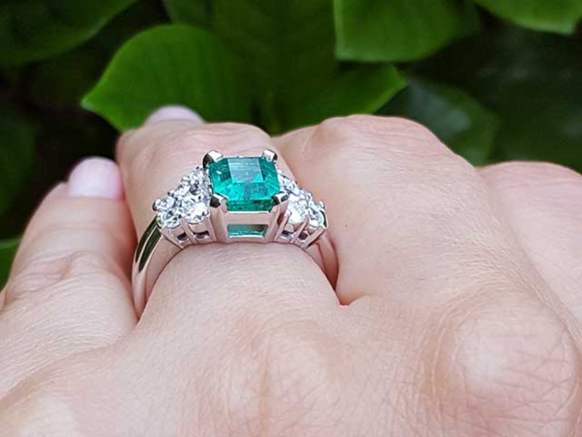 Emerald ring with diamonds and the matching band on a finger against a green leafy background.