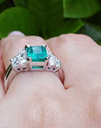 Emerald ring with diamonds and the matching band on a finger against a green leafy background.