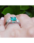 Emerald ring with diamonds and the matching band on a finger against a green leafy background.