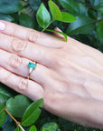 Hand wearing a ring with a natural colombian emerald against a green leafy background.